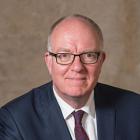 A man faces the camera. He is wearing glasses, a white shirt, red tie, and navy jacket. He is against a plain brown background.